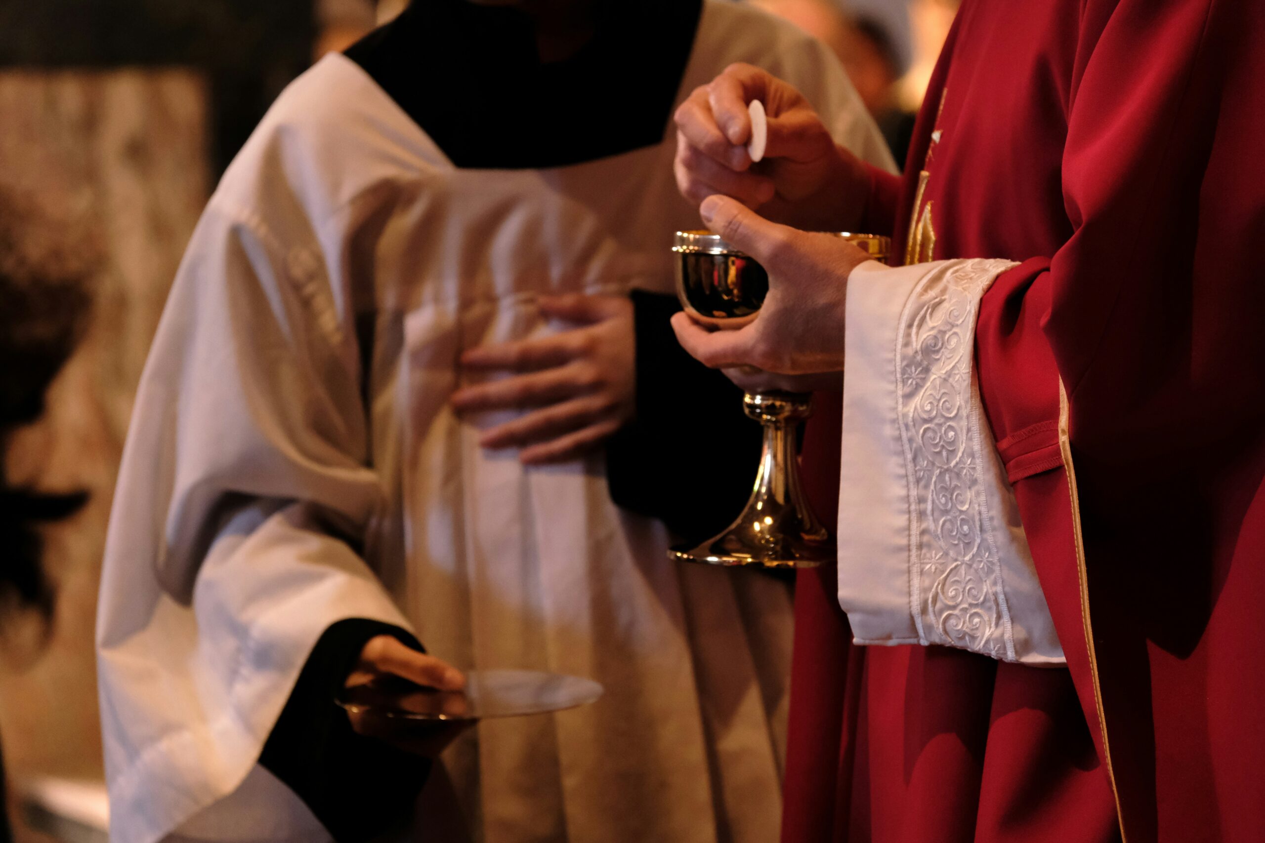 A priest in red robes holds a chalice and Eucharist wafer during a religious ceremony, assisted by an altar server in white attire, creating a solemn atmosphere. Lent, Easter, Christian, Catholic, church, mass, holy communion
