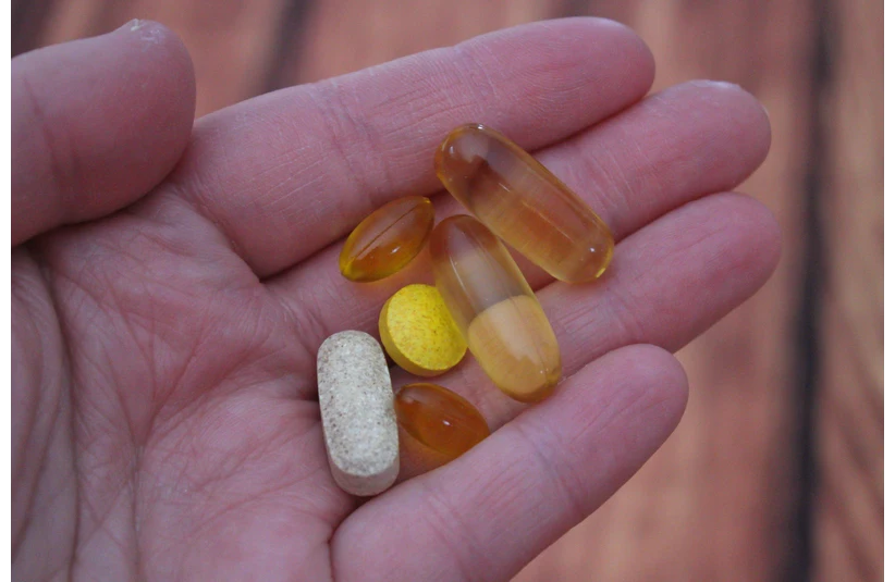 A hand holds various vitamins, including transparent softgels, a round yellow tablet, and a beige oblong pill, against a blurred wooden background.