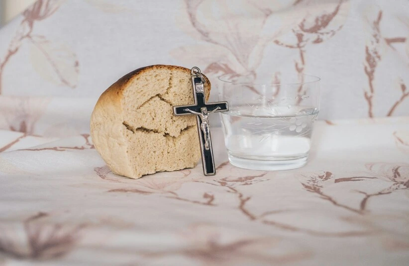 A piece of bread, a small cross, and a glass of water sit on a patterned cloth. The scene conveys simplicity and spirituality, Lent
