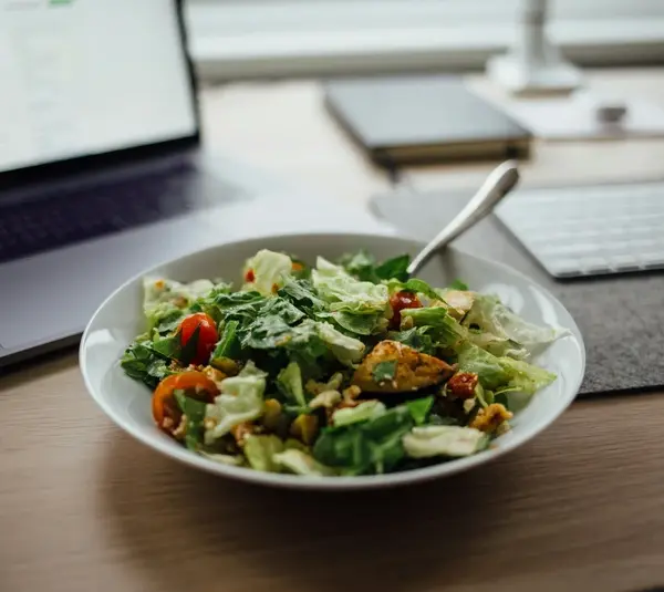 Lunch breaks for remote and hybrid workers, featuring a plate of vegetable salad next to a laptop