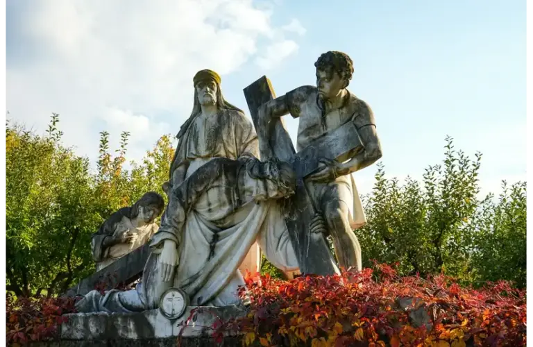 A white stone statue depicting a biblical scene of three figures supporting a cross, surrounded by greenery and vibrant red bushes under a clear sky. Ash Wednesday, Catholic, Christian, Jesus, crucifixion