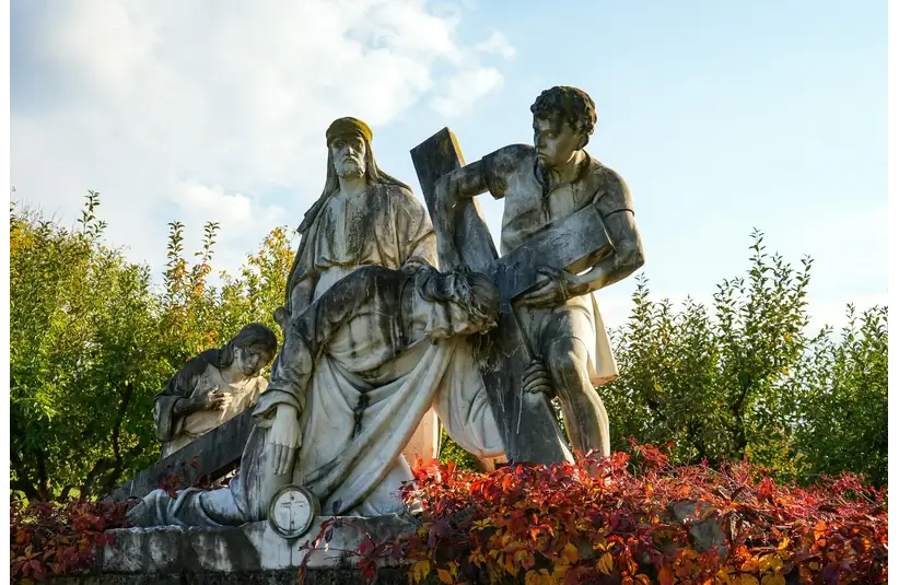 A white stone statue depicting a biblical scene of three figures supporting a cross, surrounded by greenery and vibrant red bushes under a clear sky. Ash Wednesday, Catholic, Christian, Jesus, crucifixion