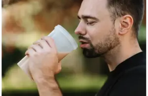 A man with a beard enjoys a protein shake from a white cup, outdoors. The background is blurred with earthy tones, creating a calm and relaxed mood. best supplements for men