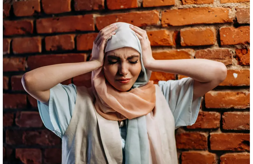 A woman in a light blue and peach hijab stands against a brick wall, holding her head with a pained expression, conveying stress or frustration.