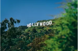 The iconic Hollywood sign sits atop a lush green hillside under a clear blue sky, framed by trees, evoking a sense of classic cinematic allure. SeeDance.