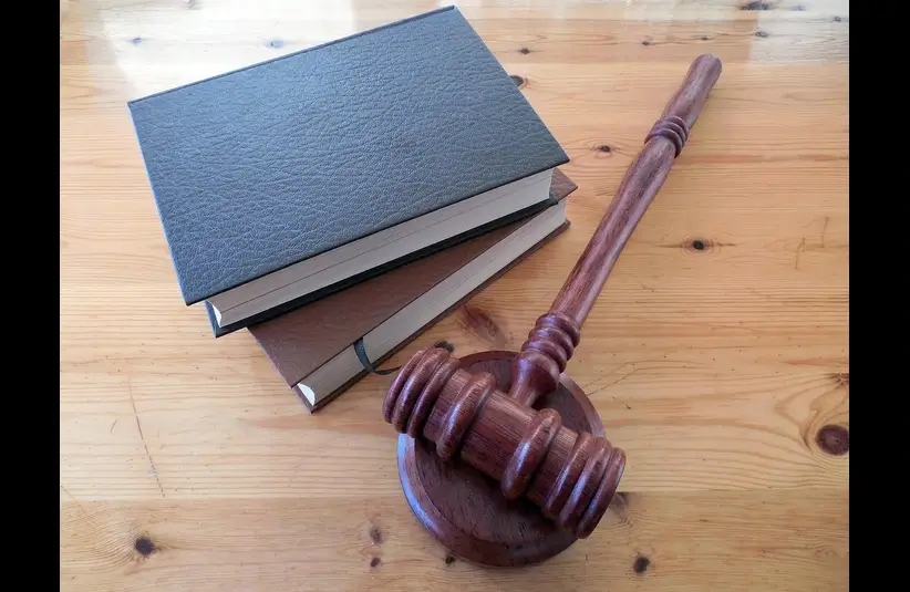 A wooden gavel rests on a wooden table beside three stacked hardcover books, symbolizing justice and law. The mood is serious and professional. Glover's suit has no court date set yet.