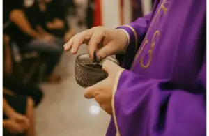 A person in a purple robe holds a small ornate bowl, possibly for a religious ritual. The setting is calm, with seated people in the background, Ash Wednesday, Lent, Catholic, Christian