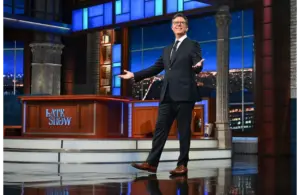 Stephen Colbert, host of The Late Show and writer for the next Lord of the Rings film, stands smiling on a brightly lit talk show stage with a cityscape backdrop. He gestures invitingly, conveying a welcoming and cheerful atmosphere.