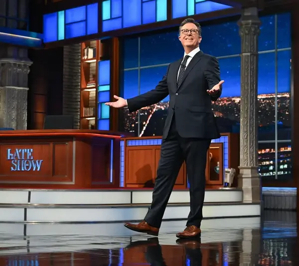Stephen Colbert, host of The Late Show and writer for the next Lord of the Rings film, stands smiling on a brightly lit talk show stage with a cityscape backdrop. He gestures invitingly, conveying a welcoming and cheerful atmosphere.