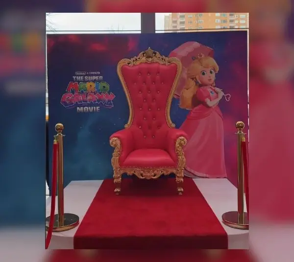 A pink plush high-back throne chair on a red carpet in front of a Super Mario Galaxy advertisement that has Princess Peach on it