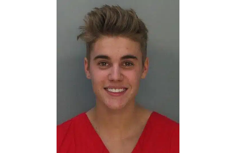 Justin Bieber's mugshot. A young person with short, styled hair wearing a red top is smiling at the camera against a plain gray background after a DUI/DWI.