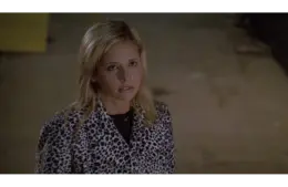 Sarah Michelle Gellar as Buffy Summers in "Buffy the Vampire Slayer" looks up with a serious expression, wearing a black and white leopard print jacket. The background is dimly lit and neutral.