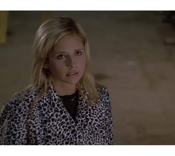 Sarah Michelle Gellar as Buffy Summers in "Buffy the Vampire Slayer" looks up with a serious expression, wearing a black and white leopard print jacket. The background is dimly lit and neutral.