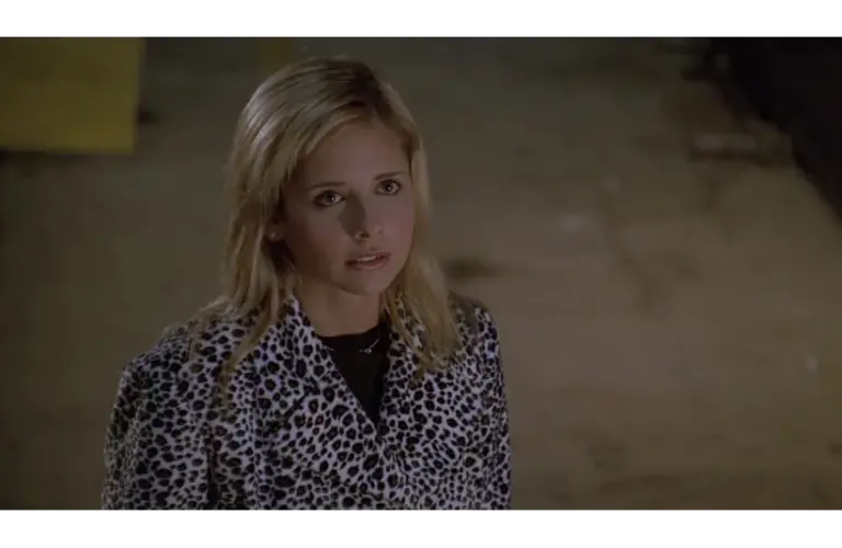 Sarah Michelle Gellar as Buffy Summers in "Buffy the Vampire Slayer" looks up with a serious expression, wearing a black and white leopard print jacket. The background is dimly lit and neutral.