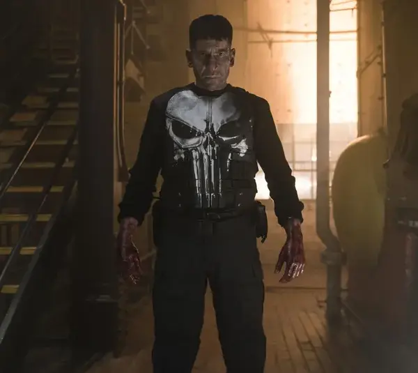 Frank Castle, aka The Punisher, played by Jon Bernthal, stands in a dimly lit industrial setting, wearing a shirt with a large white skull emblem. His expression is intense, and his hands are stained, suggesting a recent struggle. The atmosphere is tense and gritty.