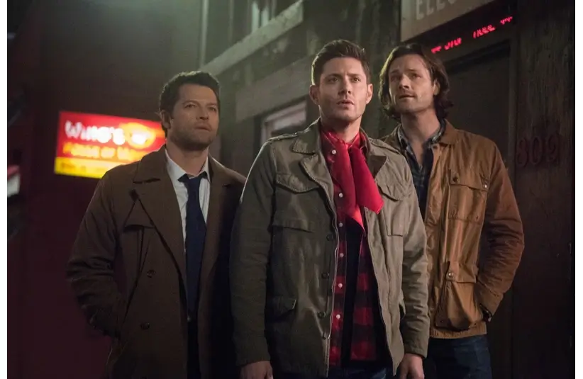 Jensen Ackles, Misha Collins, and Jared Padalecki in Supernatural, are the three men in jackets stand closely together on a dimly-lit urban street, appearing concerned. Neon signs glow in the background, adding a mysterious tone.
