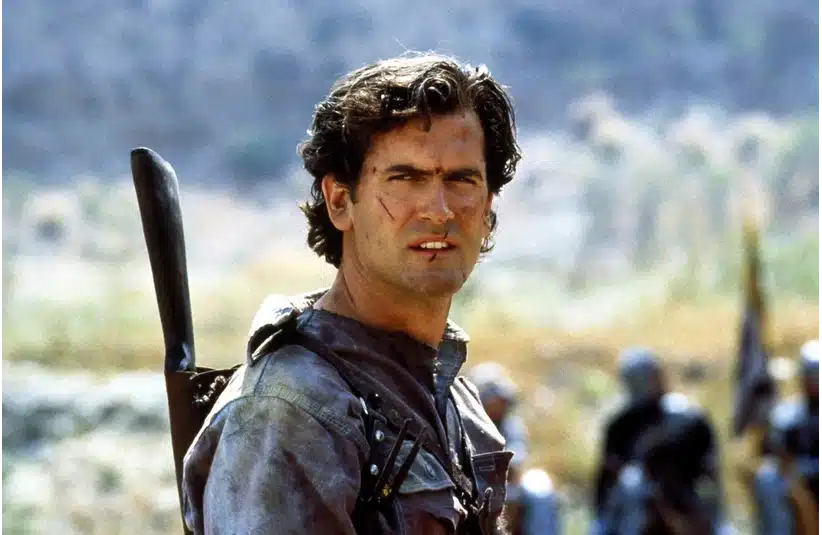 Bruce Campbell, with a rifle, stands alone in a vast field, with greenery stretching out around him.