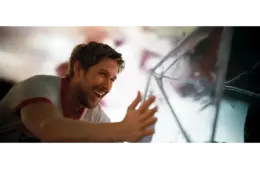 Ryan Gosling in "Project Hail Mary (2026)" smiles joyfully, reaching out towards a reflective, angular surface. The scene is brightly lit, conveying a sense of happiness and warmth.