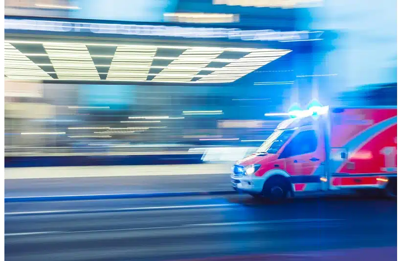 A speeding ambulance with flashing blue lights rushes down a city street. Motion blur of surrounding buildings suggests urgency and fast movement.