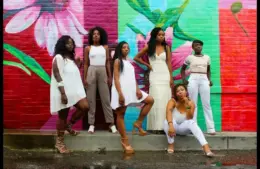 Six women pose confidently in front of a vibrant, floral mural. Their stylish white outfits contrast against the colorful red and green background. Women's History Month