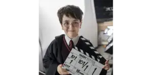 Dominic McLaughlin, as Harry Potter dressed in a wizard costume, resembling a famous fictional character, stands smiling on a film set, holding a movie clapperboard confidently.