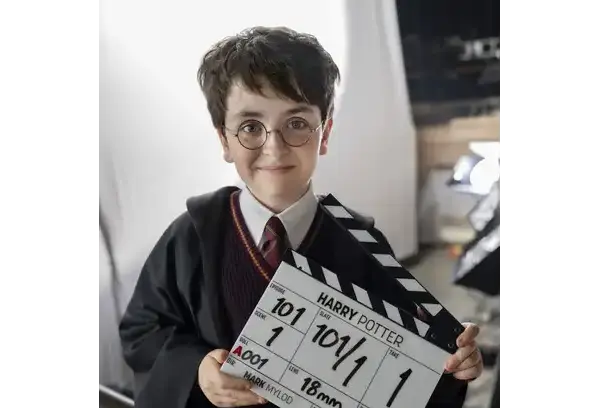 Dominic McLaughlin, as Harry Potter dressed in a wizard costume, resembling a famous fictional character, stands smiling on a film set, holding a movie clapperboard confidently.