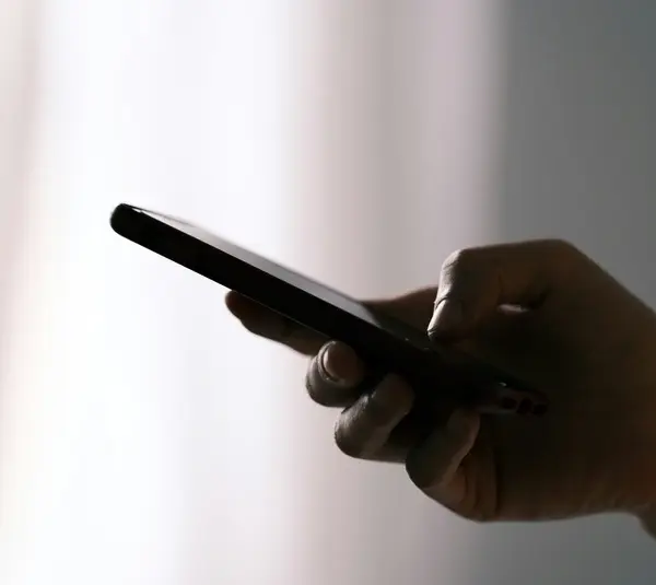 A hand holds a smartphone against a softly lit, blurred background. The image conveys a sense of focus and connectivity in a subdued, calm setting. text scam, fake recall, text message, smartphone