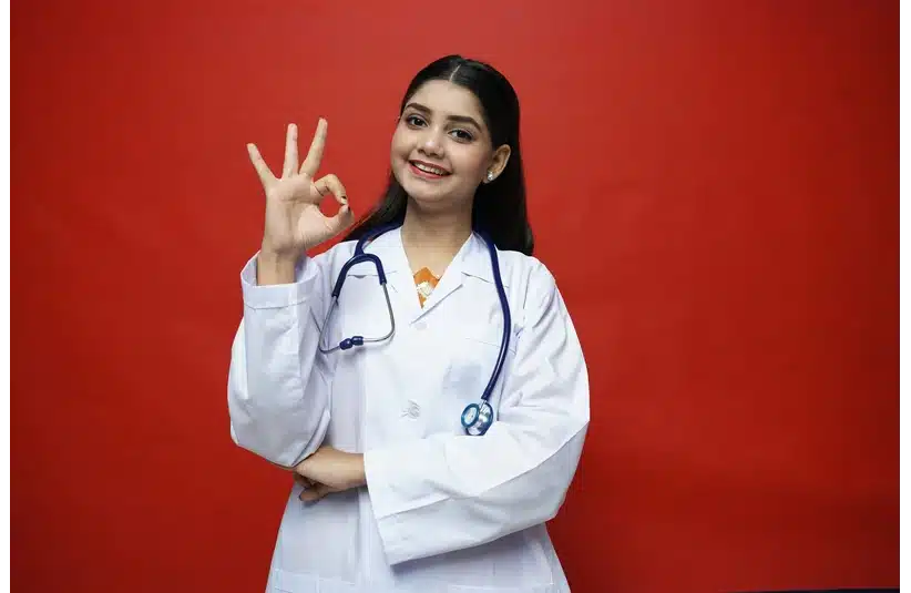 A smiling doctor in a white coat stands against a red background, making an "OK" gesture with her hand. A stethoscope hangs around her neck.