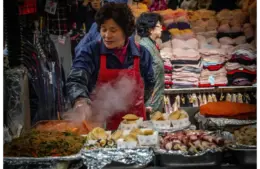 A woman wearing a red apron cooks at a bustling market stall with steaming food, surrounded by colorful textiles, conveying a lively, busy atmosphere. Women's History Month, food traditions