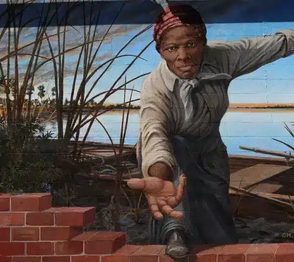 Mural of Harriet Tubman reaching out through a brick wall, set against a riverbank with reeds. The expression is determined and inviting, suggesting courage and guidance. Women's History Month