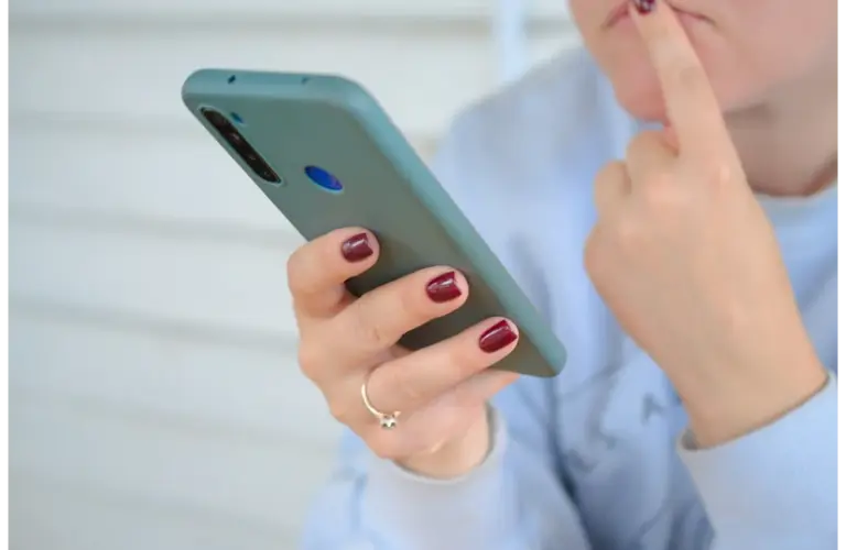 A person with maroon nail polish is holding a smartphone, looking pensive. They touch their chin with one finger, conveying thoughtfulness. text message, cell phone, thinking, text scam, social media doomscrolling