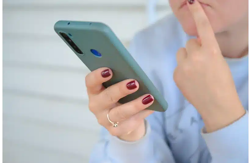 A person with maroon nail polish is holding a smartphone, looking pensive. They touch their chin with one finger, conveying thoughtfulness. text message, cell phone, thinking, text scam