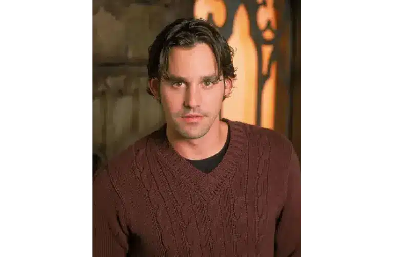 Young man, Nicholas Brendon, with dark hair in a brown cable-knit sweater stands indoors against a rustic background. He has an attentive and calm expression.