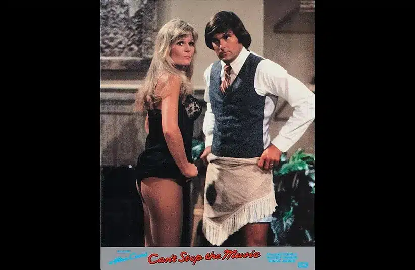 A man and woman stand side by side indoors. The woman, in a black outfit, looks confidently forward. The man wears a vest, tie, and fringe apron, posing humorously. "Can't Stop the Music" text at the bottom.
