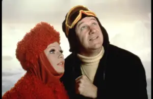 Valerie Perrine and Gene Hackman from Superman II dressed in winter clothing look upwards. The woman wears a fluffy red outfit, and the man sports ski goggles and a black jacket.
