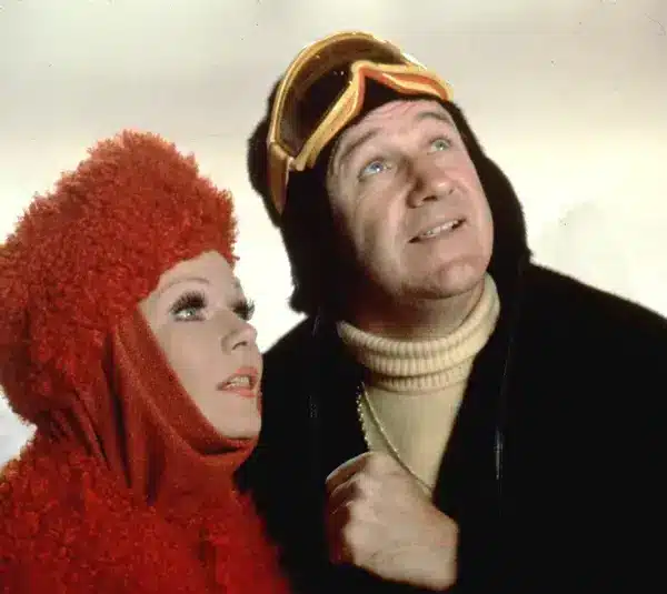 Valerie Perrine and Gene Hackman from Superman II dressed in winter clothing look upwards. The woman wears a fluffy red outfit, and the man sports ski goggles and a black jacket.