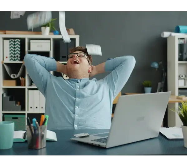 A man is celebrating success by throwing papers in the office. Lots of money falling on him in an office while sitting at a desk enjoying wealth. Happy and rich youth—a financial wellness concept.