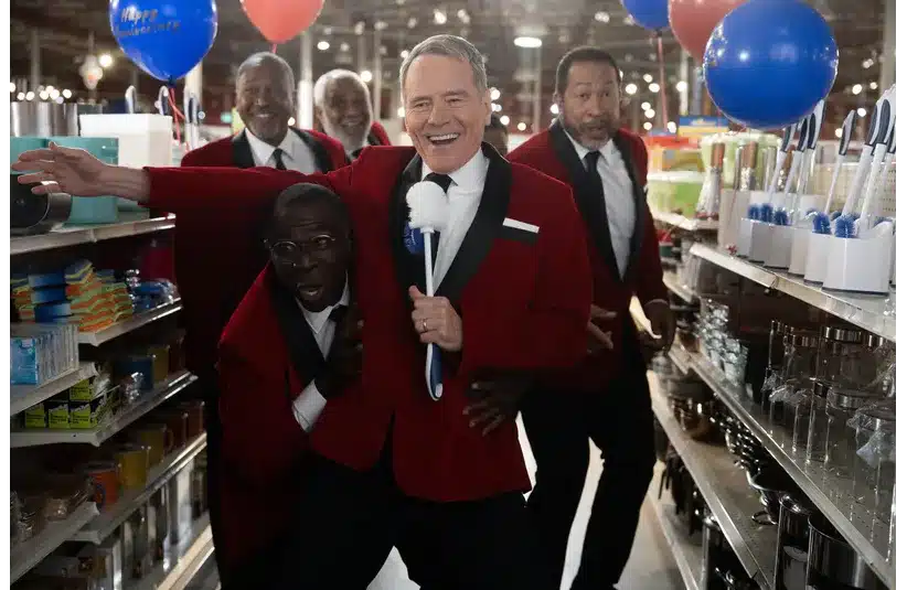 Four men in red tuxedos joyfully sing in a store aisle. One holds a white brush as a microphone. Blue and red balloons add a festive atmosphere.