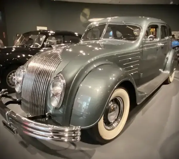 A vintage Chrysler Airflow, a car with a sleek, curved body in a glossy gray finish is displayed in a museum setting. Its design features chrome accents and whitewall tires.