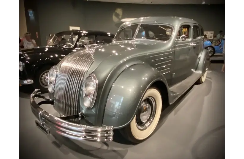 A vintage Chrysler Airflow, a car with a sleek, curved body in a glossy gray finish is displayed in a museum setting. Its design features chrome accents and whitewall tires.