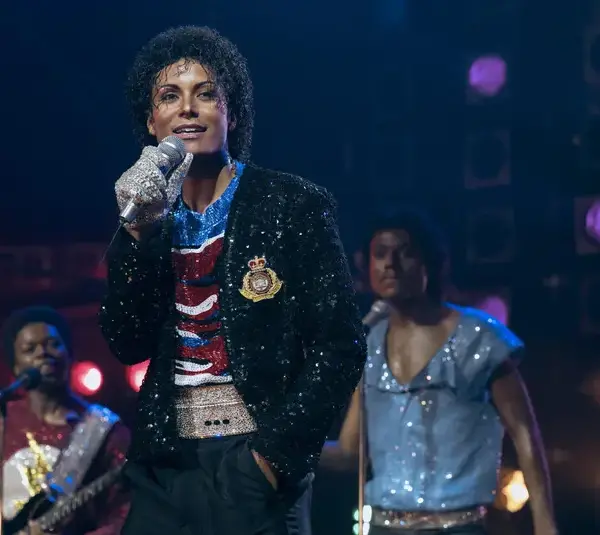 Jaafar Jackson, in "Michael (2026)" in a sequined jacket and glove, sings confidently on stage with musicians in colorful outfits. The atmosphere is vibrant and energetic.