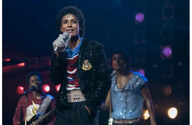 Jaafar Jackson, in "Michael (2026)" in a sequined jacket and glove, sings confidently on stage with musicians in colorful outfits. The atmosphere is vibrant and energetic.