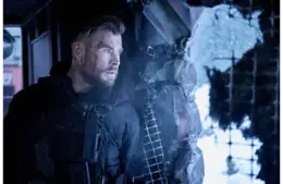 Chris Hemsworth, returning for Extraction 3, in tactical gear, stands tensely by a broken window, looking out with intensity. The scene is cold and war-torn, evoking suspense.