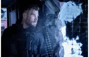 Chris Hemsworth, returning for Extraction 3, in tactical gear, stands tensely by a broken window, looking out with intensity. The scene is cold and war-torn, evoking suspense.