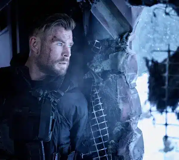 Chris Hemsworth, returning for Extraction 3, in tactical gear, stands tensely by a broken window, looking out with intensity. The scene is cold and war-torn, evoking suspense.