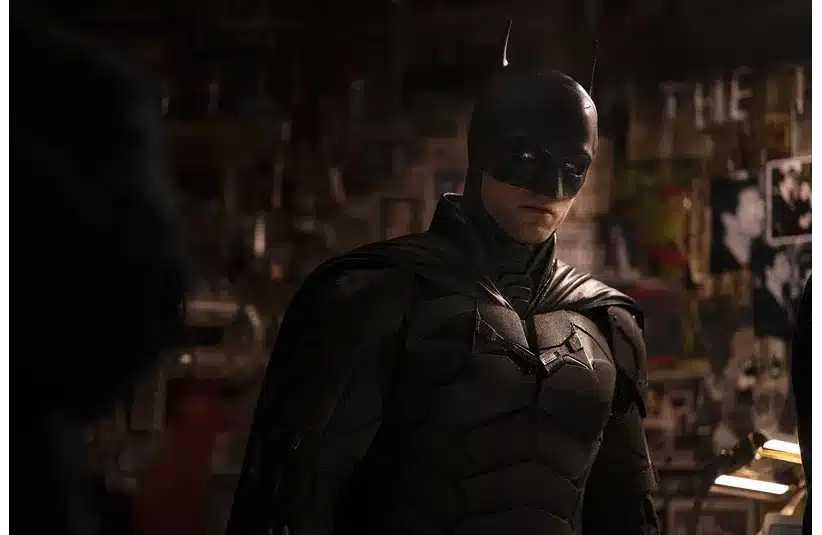 Batman, a person in a dark, armored costume with a mask and pointed ears stands in a dimly lit room with photos and newspaper clippings on the wall, conveying a mysterious and intense atmosphere.