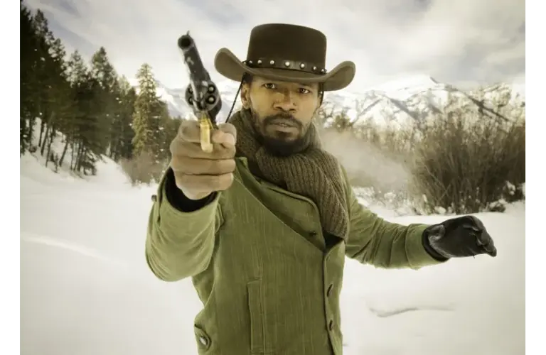 Django, played by Jamie Foxx, in a green coat and brown hat, aims a revolver intently in a snowy forest with distant mountains. The scene conveys tension and determination.