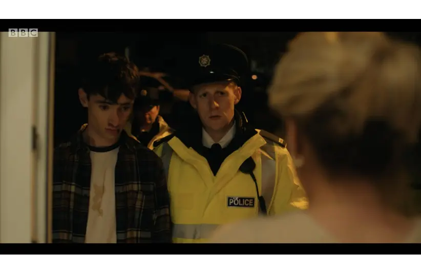 Michael Patrick MND -A young man in a plaid shirt stands at a doorway next to a police officer in a yellow jacket. The scene is tense and dimly lit, conveying urgency.
