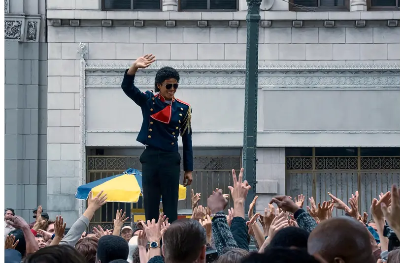 Jaafar Jackson in Michael (2026), in a military-style jacket, waves from a platform above an excited crowd reaching up. The scene is set against an urban backdrop with a lively, celebratory tone.
