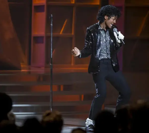 Jaafar Jackson in "Michael (2026)" performs on stage in a sparkling black jacket and glove, singing passionately into a microphone. The audience watches intently, creating an energetic atmosphere.